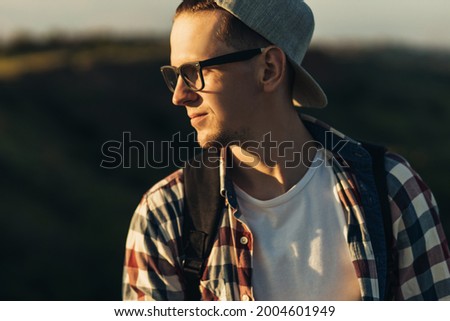 Portrait of a young handsome man, wearing glasses and a cap, smiling young man, standing on the street in nature, against the background of a beautiful forest