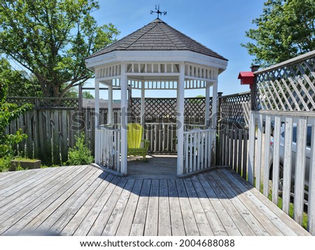 A beautiful gazebo tucked into the corner of a backyard, deck made of wood. The deck is surrounded with a wooden gate with a lattice design. There is a weathervane on top of the gazebo and a birdhouse Royalty-Free Stock Photo #2004688088