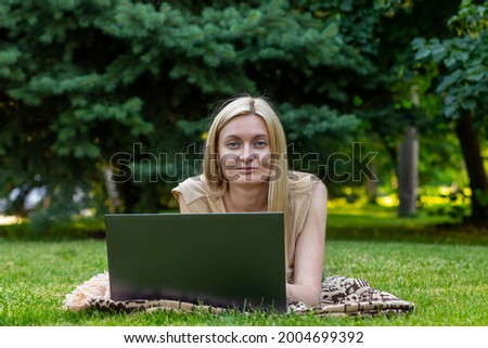 young woman with laptop outside lies on the lawn. she looks at the camera and smiles