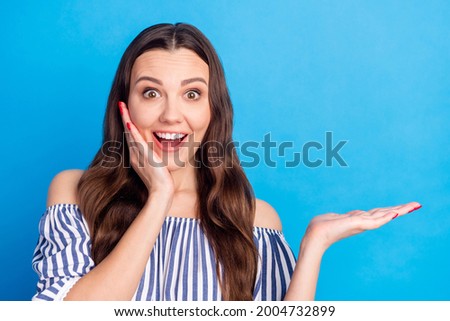 Photo portrait woman showing copyspace keeping on palm smiling amazed touching cheek isolated vivid blue color background
