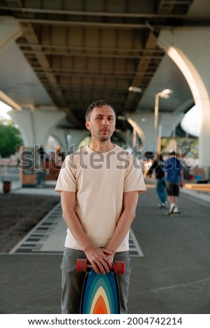 Millennial man holding bright longboard stand in urban skate park with skateboarders practice on background. Portrait of young adult hipster longboarder before exercising and skating in city skatepark