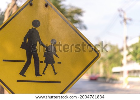 The close up shot of yellow notice sign of school and people crossing the street at crosswalk with beautiful street bokeh as a background during bright sunny day