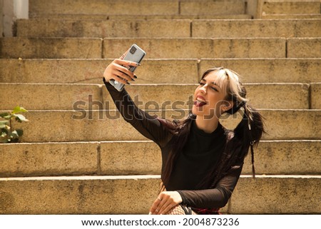Young and beautiful girl with pigtails and punk style sitting on some stairs taking a selfie with her mobile phone while sticking out her tongue.