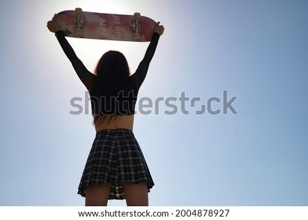Young girl in punk style with black blouse and plaid skirt raising a skateboard above her head looking backwards facing the sun.