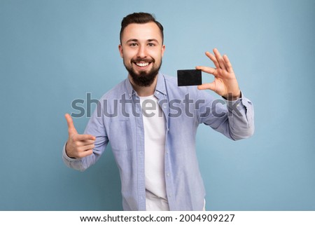 photo shot of Handsome smiling brunette bearded young man wearing stylish blue shirt and white t-shirt isolated over blue background wall holding credit card looking at camera