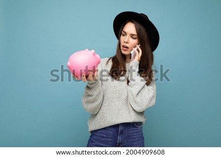 Photo of asking serious thoughtful young attractive pretty woman with dark streight long hair wearing casual grey sweater and black hat isolated over blue background with free space, holds pink pig