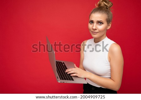 Side profile photo shot of beautiful asking amazed surprised astonished blond young lady with gathered hair wearing white t-shirt holding computer laptop typing on keyboard looking at camera isolated