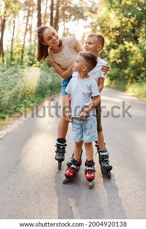 Happy smiling woman roller skating with her children in summer park, mother looking at kids with toothy smile, family rollerblading and having fun together.