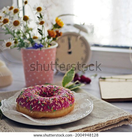 Summer breakfast with a cup of coffee and a donut in pink glaze on a background of wild flowers of chamomiles, fresh berries, an alarm clock on a wooden table.Home cozy summer rustic background
