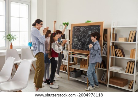 Happy teacher and children playing games and having fun in class. Cheerful school students standing in front of classroom board and applauding classmate for good creative interesting presentation