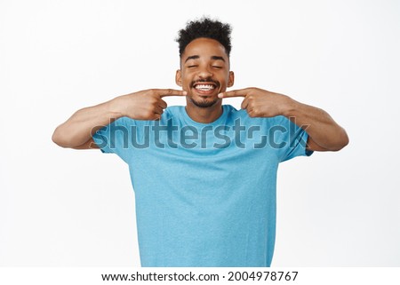 Dental clinic, advertising concept. Happy african american guy pointing fingers at perfect white smile, smiling and showing it, point at mouth, standing cheerful against studio background