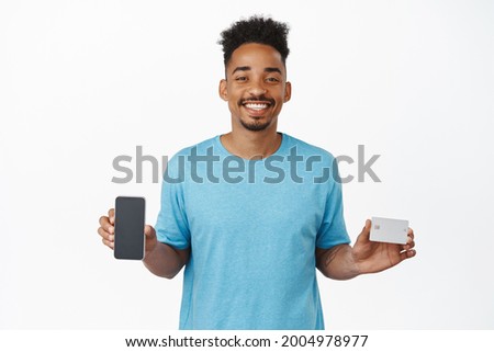 Happy smiling african american guy showing credit card and mobile phone screen, shopping on smartphone, recommending internet store, standing against white background