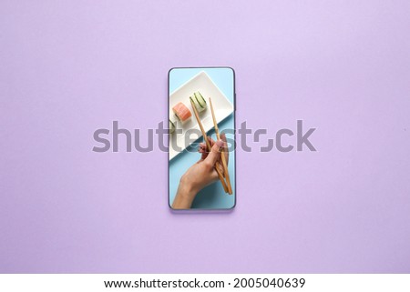 Female hand with chopsticks and sushi rolls on screen of mobile phone on color background