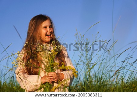 Portrait of cute smiling caucasian girl walking in the meadow holding a bouquet of wildflowers against of clear blue sky.  Summer sunset. Happy Childhood concept.                                