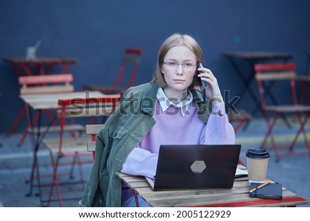 Young woman freelancer in glasses sits on cafe terrace at table with black laptop and types under bright sunlight. Female student distant learning foreign languages and working distantly. Royalty-Free Stock Photo #2005122929