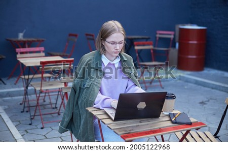 Young woman freelancer in glasses sits on cafe terrace at table with black laptop and types under bright sunlight. Female student distant learning foreign languages and working distantly. Royalty-Free Stock Photo #2005122968