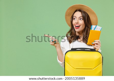 Close up excited tourist woman in casual clothes hat hold passport ticket suitcase point finger aside workspace isolated on green background Passenger travel abroad weekend getaway Air flight concept Royalty-Free Stock Photo #2005135889