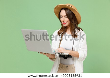 Traveler fun smiling tourist woman in casual clothes hat camera hold laptop pc computer chat online isolated on green background Passenger travel abroad on weekends getaway. Air flight journey concept