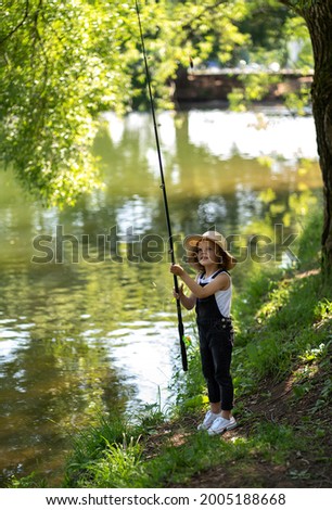 A little girl with curly hair in a straw hat and black overalls stands with a fishing rod by the pond. Child fishing on a summer day