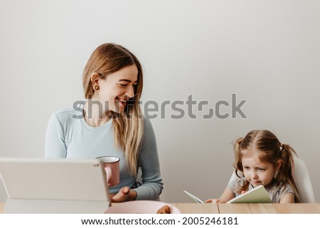 blonde woman in glasses sitting at a table with a computer tablet, eating chocolate chip cookies and working remotely, drinking coffee or tea. girl with mom reads a book at the table