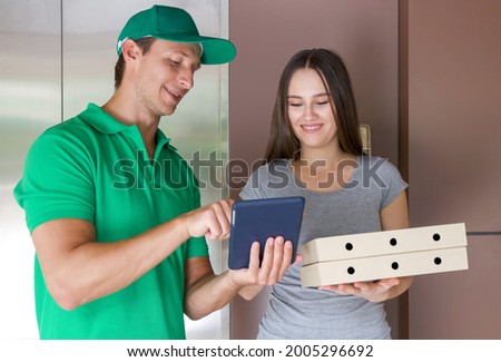 The delivery staff in a green uniform allows the customer to sign to receive the product with application on tablet computer after delivering pizza boxes. Food and parcels delivery concept.