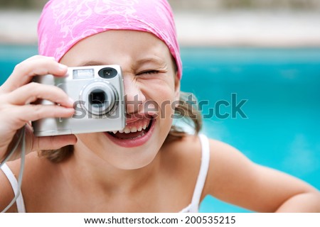 Close up portrait of a young girl using a digital photo camera to take pictures pointing at the camera with a blue water swimming pool space in the background. Active children learning, outdoors.