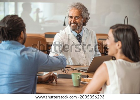 Doctor speaking to young couple. Doctor Using Tablet Computer Discussing Treatment With Patients. A photo of happy male doctor shaking hands with patient. Mature medical professional is in clinic. 