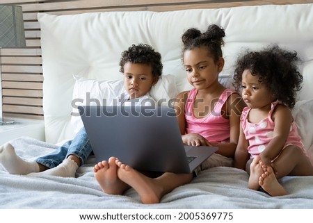Cute african american family of children sister and brother watching online cartoons on laptop before bedtime. Little toddler boy and preschooler girls sitting on bed using pc sitting together at home