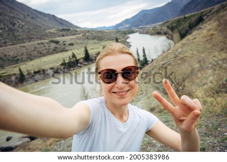 Happy young adult woman in sunglasses and white T-shirt takes a selfie, laughing into a smartphone camera against the mountain river landscape. Travel, blogging, active lifestyle.