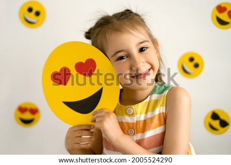  A  girl, smiling with all her teeth, holds a love emoji in her hands. World emoji day. Anthropomorphic smile Face.  Emotions.
