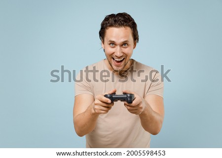 Young smiling happy excited caucasian fun man 20s wearing casual basic beige t-shirt play pc game with joystick console isolated on pastel blue background studio portrait. People lifestyle concept.