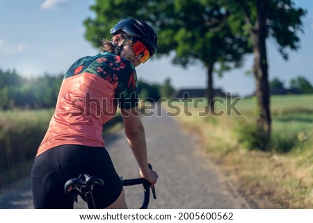 Portrait of a young cyclist, from behind, standing on her bike, looking back at the camera, with a confident expression, ready to start her outdoor cycling training. Gender equality in sport concept Royalty-Free Stock Photo #2005600562