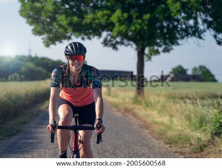 Frontal photo of a young woman cyclist, smiling, riding her road bike, in the middle of nature, illuminated by sunlight. She is wearing a helmet, sunglasses, and a pink cycling outfit. Royalty-Free Stock Photo #2005600568