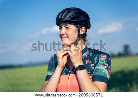 Close up outdoor portrait of a young woman cyclist, illuminated by sunlight, putting on her helmet before starting her cycling training. She grimaces with her eyes because of the sunlight Royalty-Free Stock Photo #2005600571