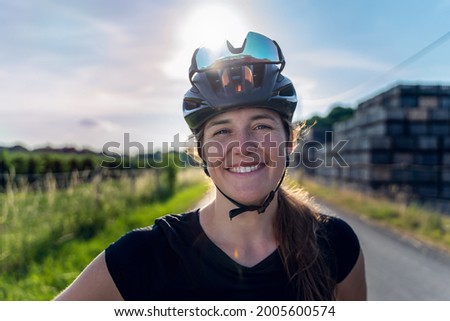 Outdoor Back light portrait, of a woman cyclist, with sunglasses placed backwards in her helmet. She looking at the camera with serene and satisfied expression. Royalty-Free Stock Photo #2005600574