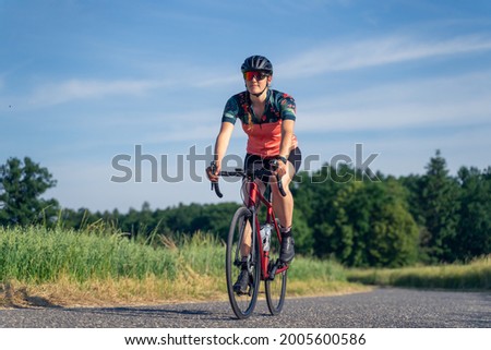 Angled photo, of a young woman professional cyclist, riding her road bike, on a paved road amidst nature, illuminated by sunlight. Sport Equality concept. Royalty-Free Stock Photo #2005600586