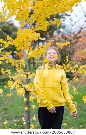 A teenager in a yellow sweater against a background of yellow leaves. The girl throws up the leaves. The girl rejoices in the autumn. Vertical frame.