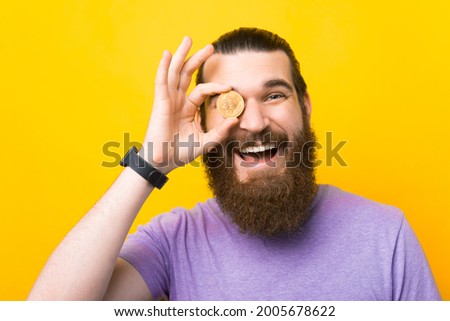 Cheerful bearded young man covering eye with bitcoin coin currency on yellow colour background studio portrait.