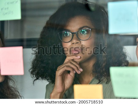 Young thoughtful African American girl student employee leader at modern office university campus looking at adhesive sticky notes on glass planning project strategy. Shot through glass reflection. Royalty-Free Stock Photo #2005718510
