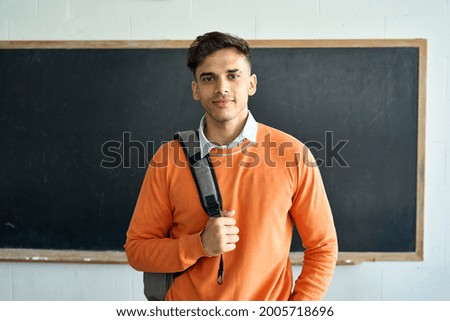 Portrait of young happy confident Indian latin Hispanic high school college university student standing in classroom on black board background, holding backpack on shoulder looking at camera.