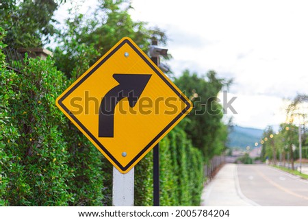 There is a sign indicating a right turn on the roadside and there is a green wall of trees. Royalty-Free Stock Photo #2005784204