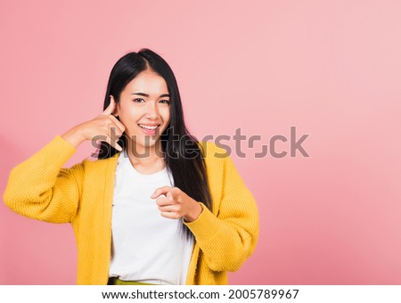 Portrait Asian beautiful young woman smiling doing phone gesture with hand fingers on telephone and pointing to camera while making call back to me gesture, studio shot isolated pink background