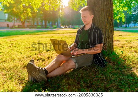 A young man is sitting under a tree and working with a laptop in a green summer park at sunset