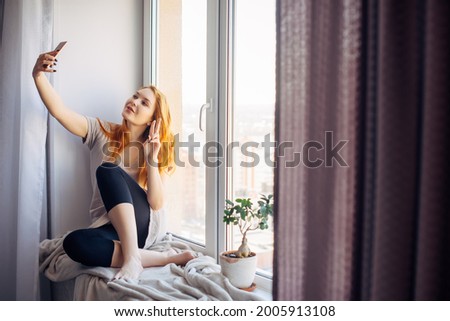 Pretty young woman with long red hair takes a selfie on her smartphone while sitting on the windowsill. Beautiful girl with a phone in a home environment. Technology and social networks concept.