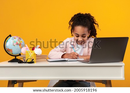 Remote Education Concept. Smiling young black teenage girl sitting at table, using laptop and writing in her notebook or diary. African schoolgirl watching online course, taking notes, doing homework