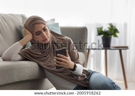 Woman upset after reading bad message on phone at home. Frustrated, sad, suffering, thinking, depressed, stressed, unhappy arab woman in hijab look at smartphone in living room interior on floor