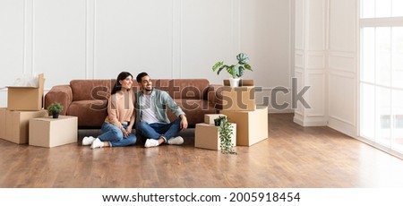 Moving Day. Lovely couple sitting on the floor in new home with cardboard boxes around. Happy man and woman dreaming, looking aside at window, free copy space, panoramic banner for advertisement