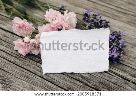 Floral stationery still life scene. Blank greeting card mock-up on old wooden table background with lavender and little pink roses flowers. Top view, selective focus. Summer birthday, wedding concept