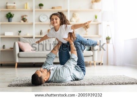 Adorable little curly girl spending time with her daddy at home, middle-eastern young man father playing airplane with his cute daughter, laying on floor in living room, lifting kid up, copy space Royalty-Free Stock Photo #2005939232