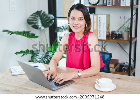 A cheerful middle-aged Asian businesswoman in relaxed casual dress working at home, checking email on computer laptop, writing on financial accounting document paper. Business stock photo